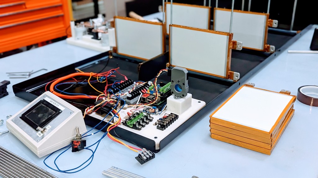 Prototype battery pack assembly on an engineering workbench: BMS monitor, color-coded wiring, terminal blocks, and stacked cells in Kapton tape.