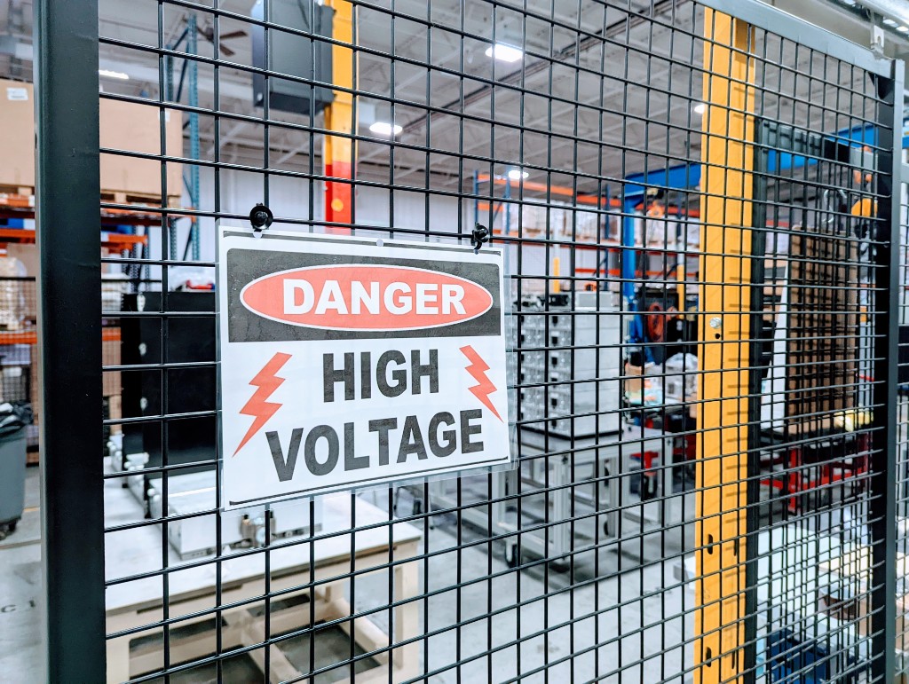 High-voltage test area behind safety mesh, with warning signage and battery engineering equipment.