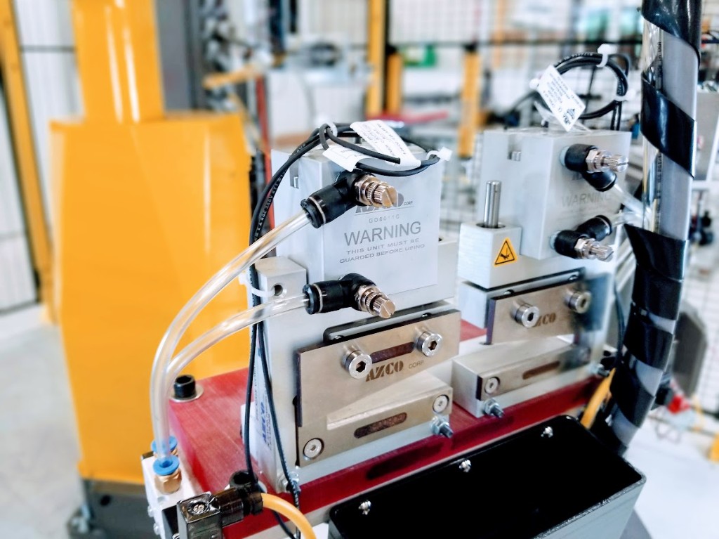 Precision in-line feeding units on a red machine base with pneumatic tubing, harnessed cabling, and safety labeling; yellow industrial robot structure and wire-mesh guarding visible in the background.