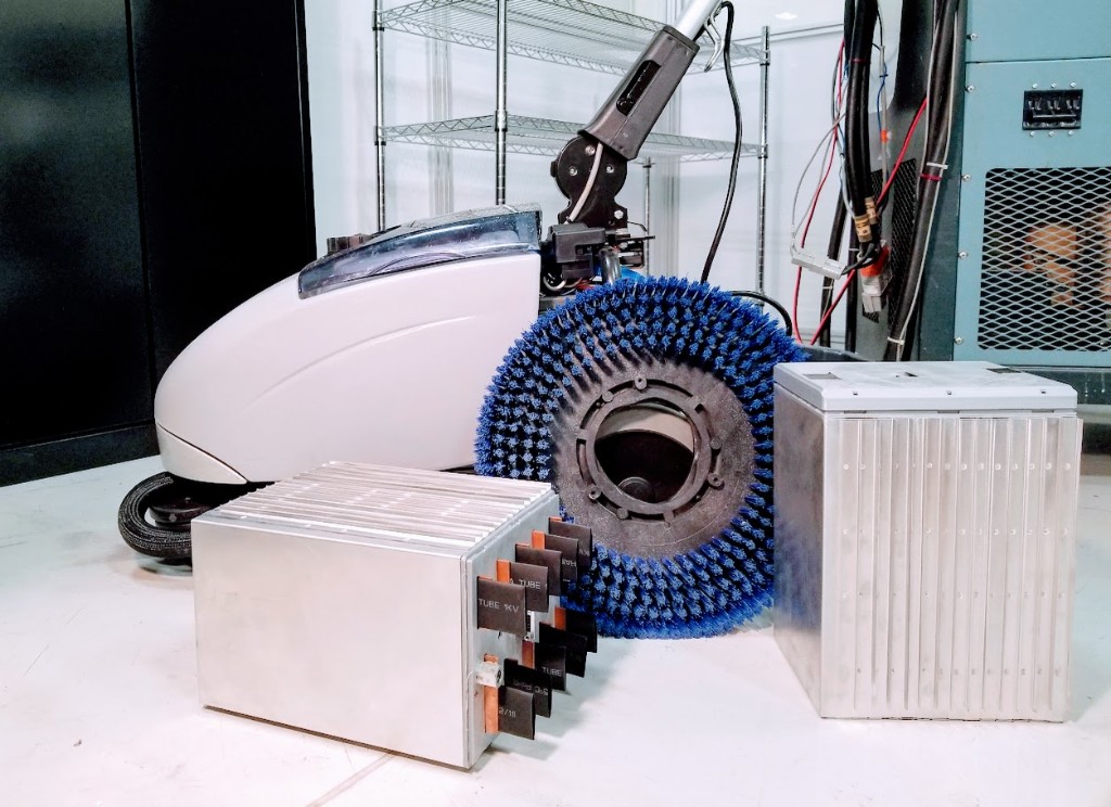 Two modular aluminum battery enclosures with insulated copper busbar tabs on a light floor, next to an industrial floor scrubber with a large blue brush; wire shelving and a teal equipment cabinet in the background.